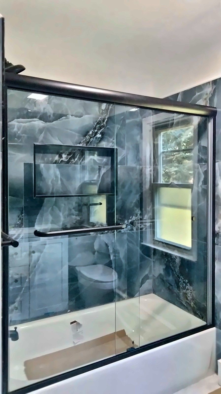 Tub with black-framed sliding glass doors and blue marble-look tile with a recessed niche