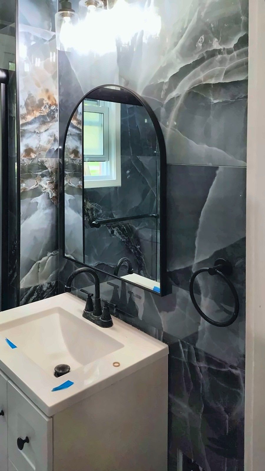 White vanity with an arched black mirror and vanity light on blue marble-look tile